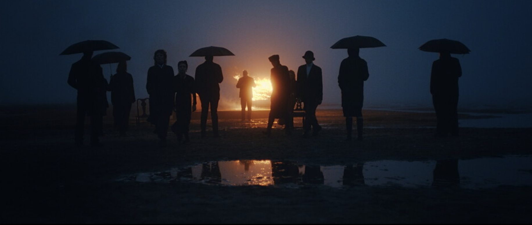 Mourners under umbrellas walking past a fire on a dark shoreline
